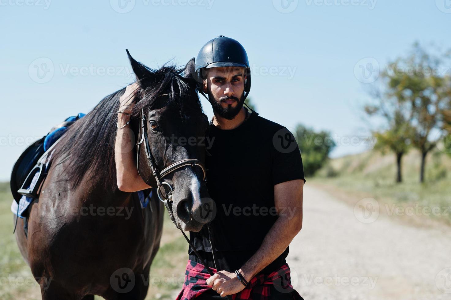 El hombre árabe de barba alta usa casco negro con caballo árabe. foto