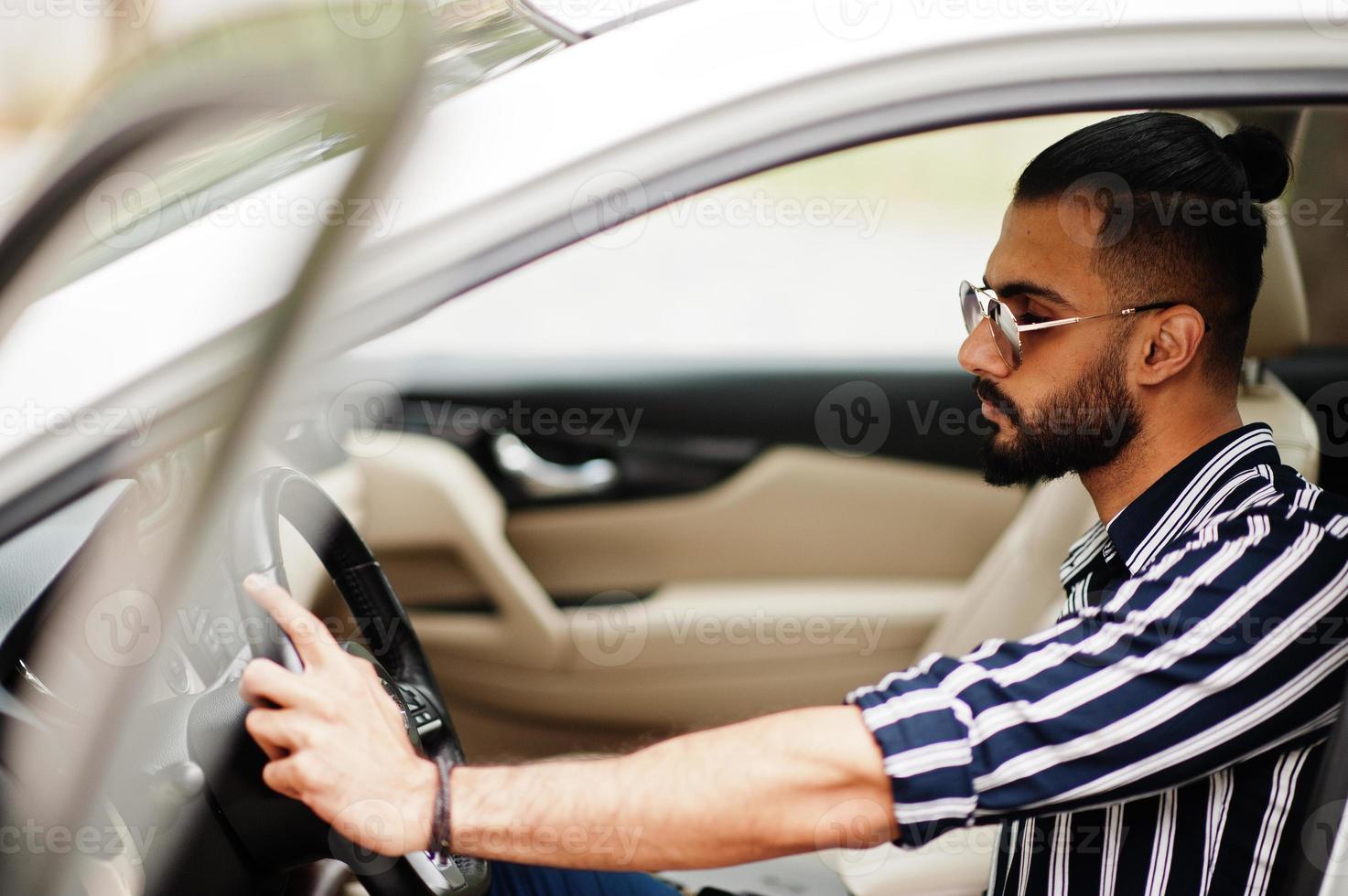 Successful arab man wear in striped shirt and sunglasses pose behind the wheel of  his white suv car. Stylish arabian men in transport. photo