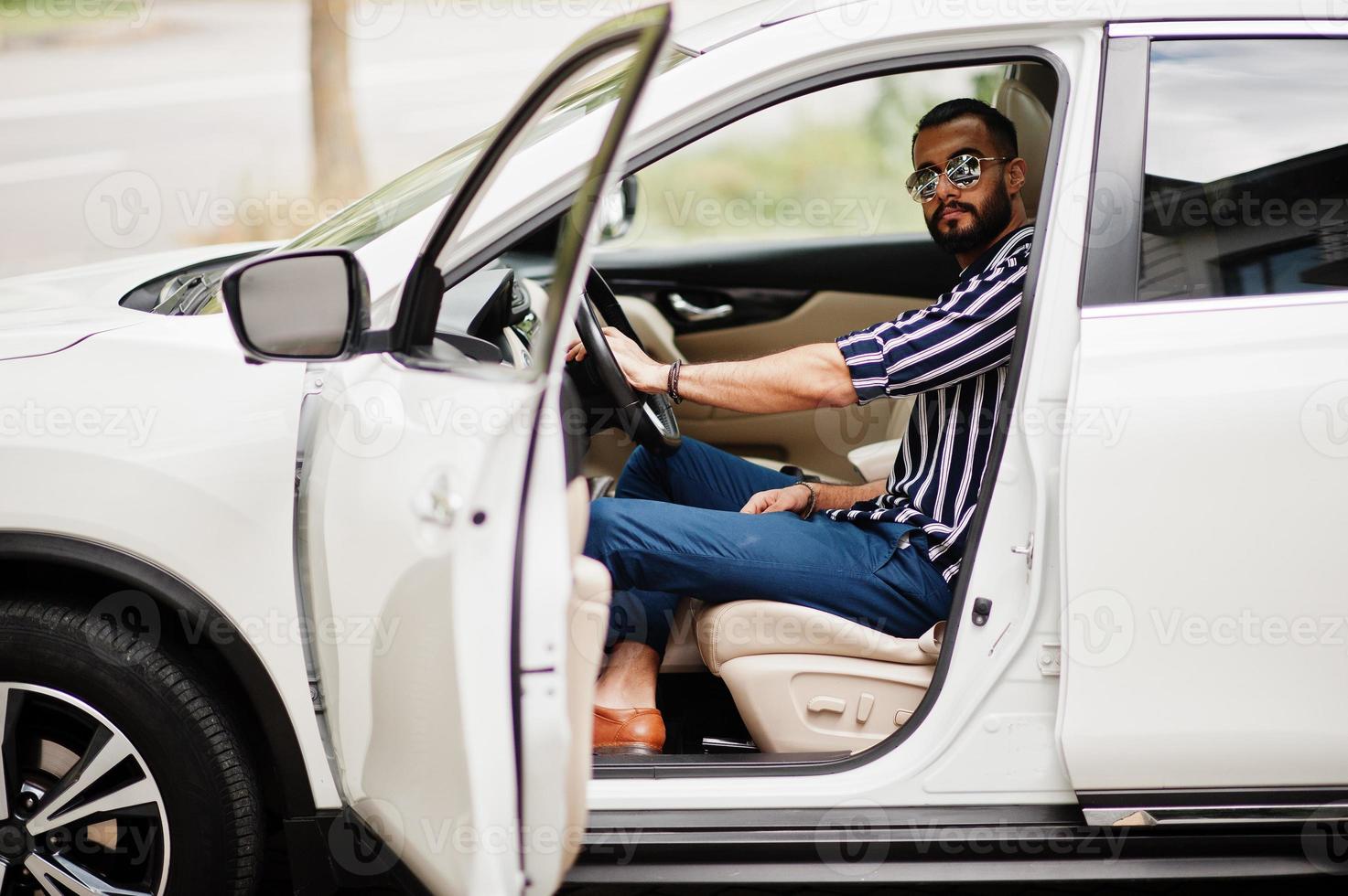 Successful arab man wear in striped shirt and sunglasses pose behind the wheel of  his white suv car. Stylish arabian men in transport. photo