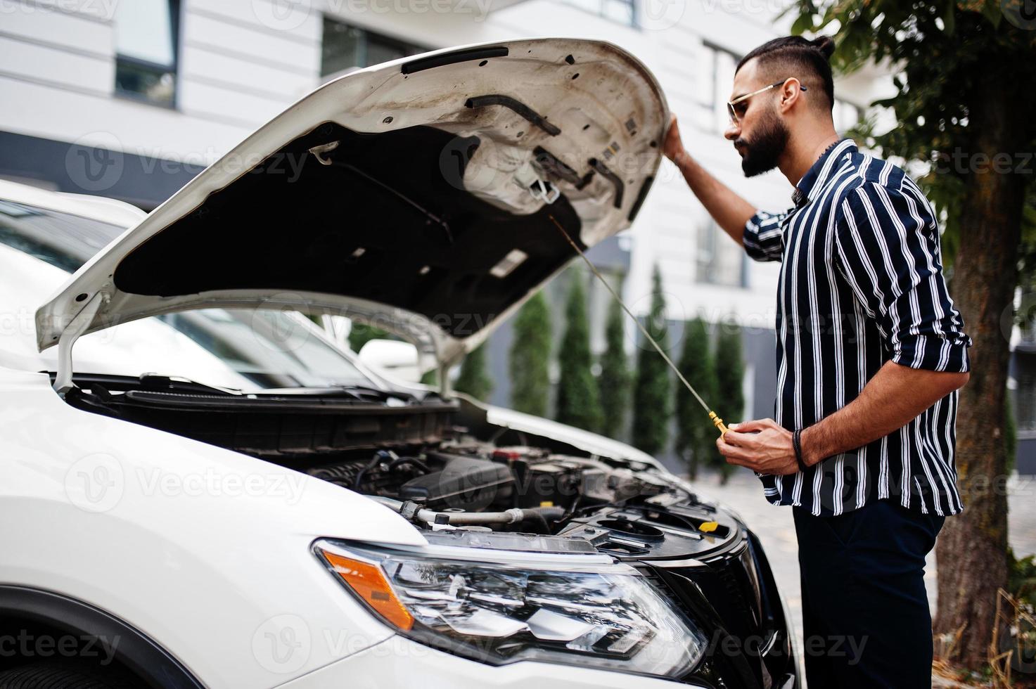 Successful arab man wear in striped shirt and sunglasses pose near his white suv car, check engine with open hood. photo