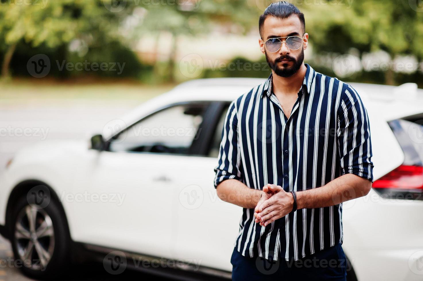 Successful arab man wear in striped shirt and sunglasses pose near his white suv car. Stylish arabian men in transport. photo