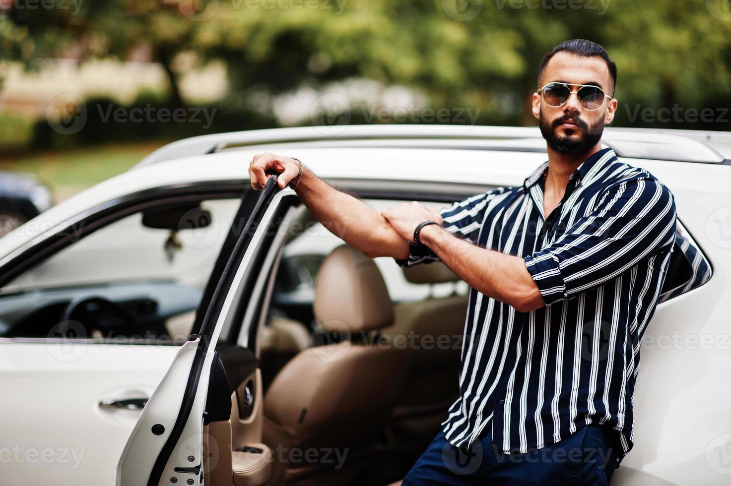 Successful arab man wear in striped shirt and sunglasses pose near his white suv car. Stylish arabian men in transport. photo