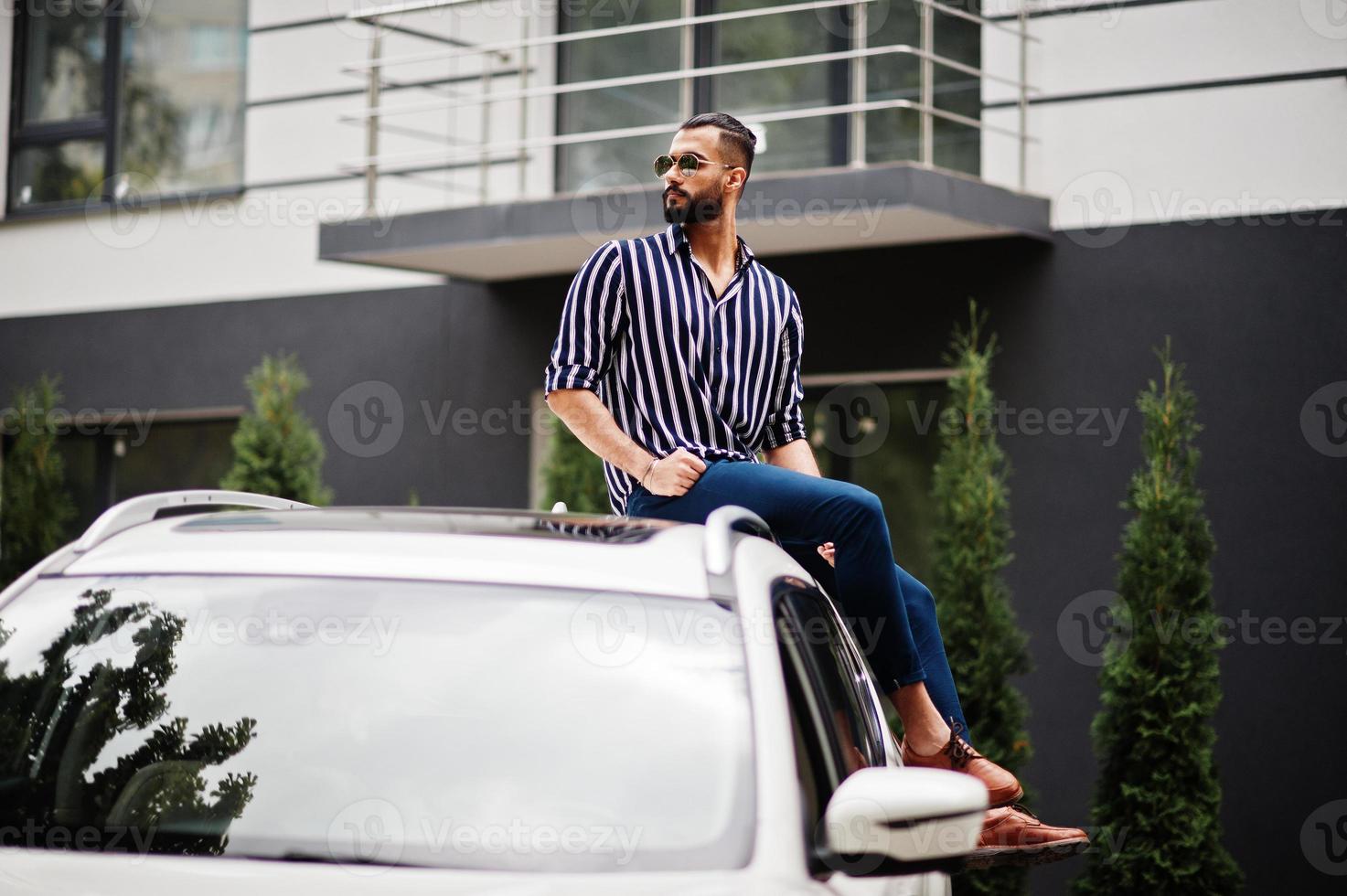 Successful arab man wear in striped shirt and sunglasses sitting on the roof of his white suv car. photo