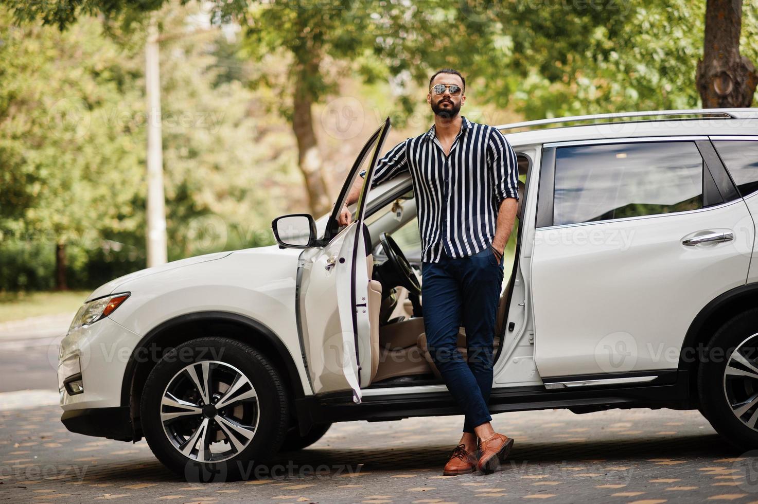 Successful arab man wear in striped shirt and sunglasses pose near his white suv car. Stylish arabian men in transport. photo