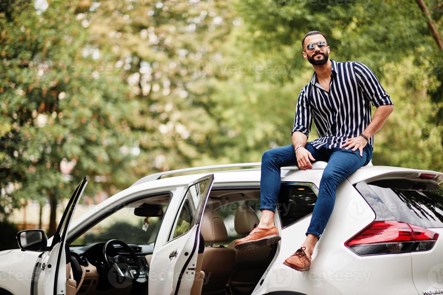 Successful arab man wear in striped shirt and sunglasses sitting on the roof of his white suv car. photo