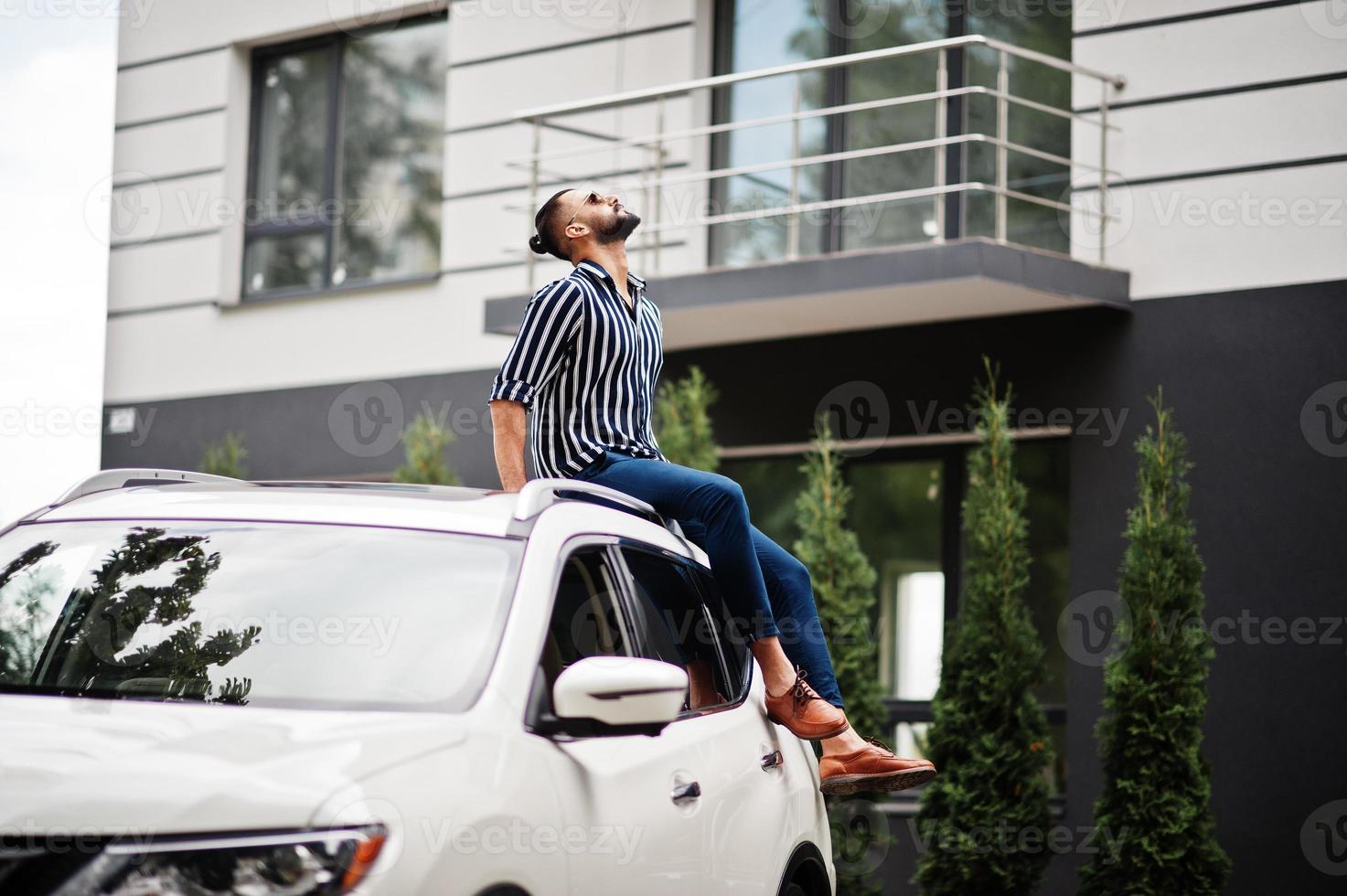 Successful arab man wear in striped shirt and sunglasses sitting on the roof of his white suv car. photo