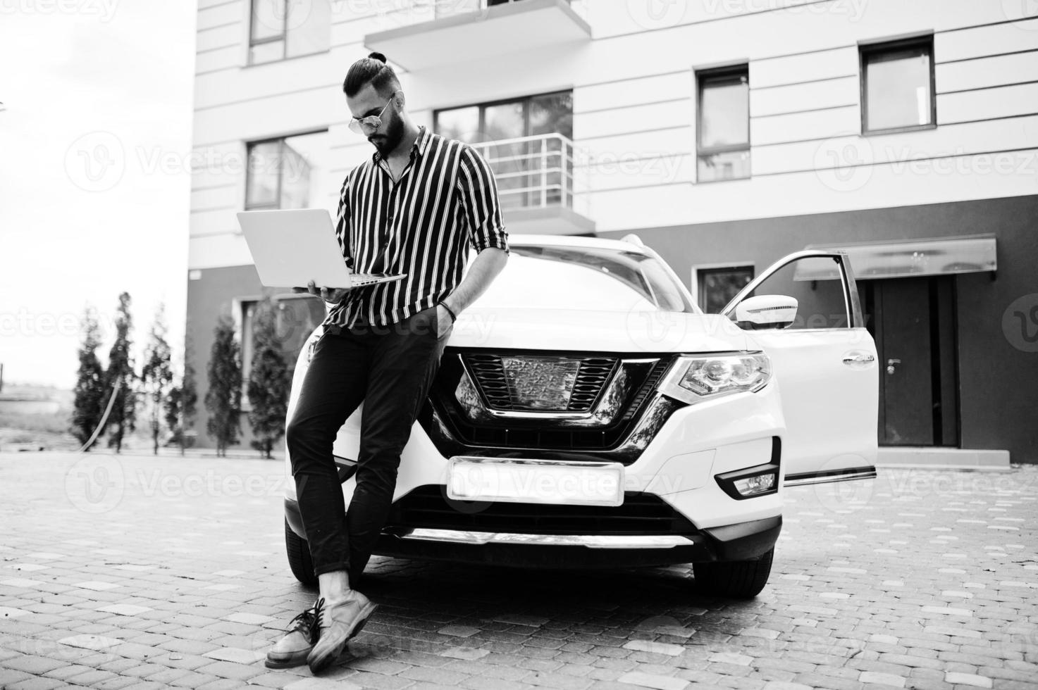 Successful arab man wear in striped shirt and sunglasses pose near his white suv car with laptop in hand. photo