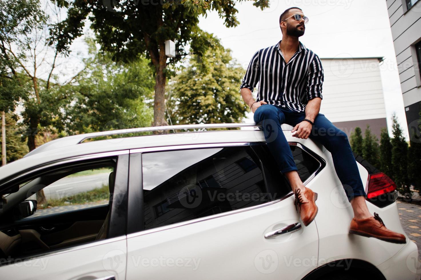 Successful arab man wear in striped shirt and sunglasses sitting on the roof of his white suv car. photo