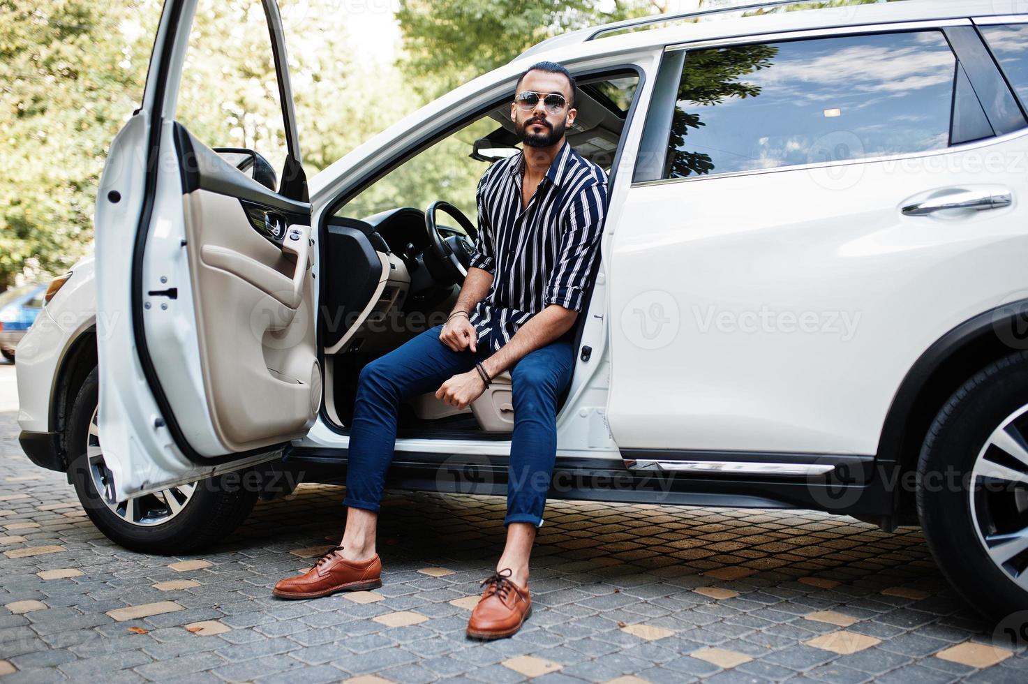 Successful arab man wear in striped shirt and sunglasses pose behind the wheel of  his white suv car. Stylish arabian men in transport. photo