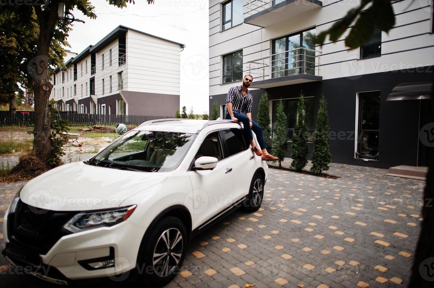 Successful arab man wear in striped shirt and sunglasses sitting on the roof of his white suv car. photo