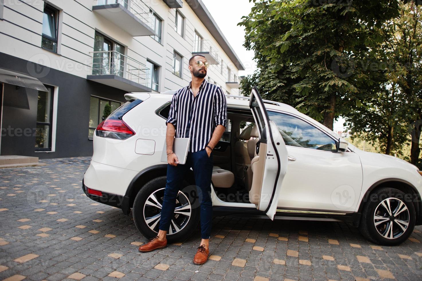 Successful arab man wear in striped shirt and sunglasses pose near his white suv car with laptop in hands. photo
