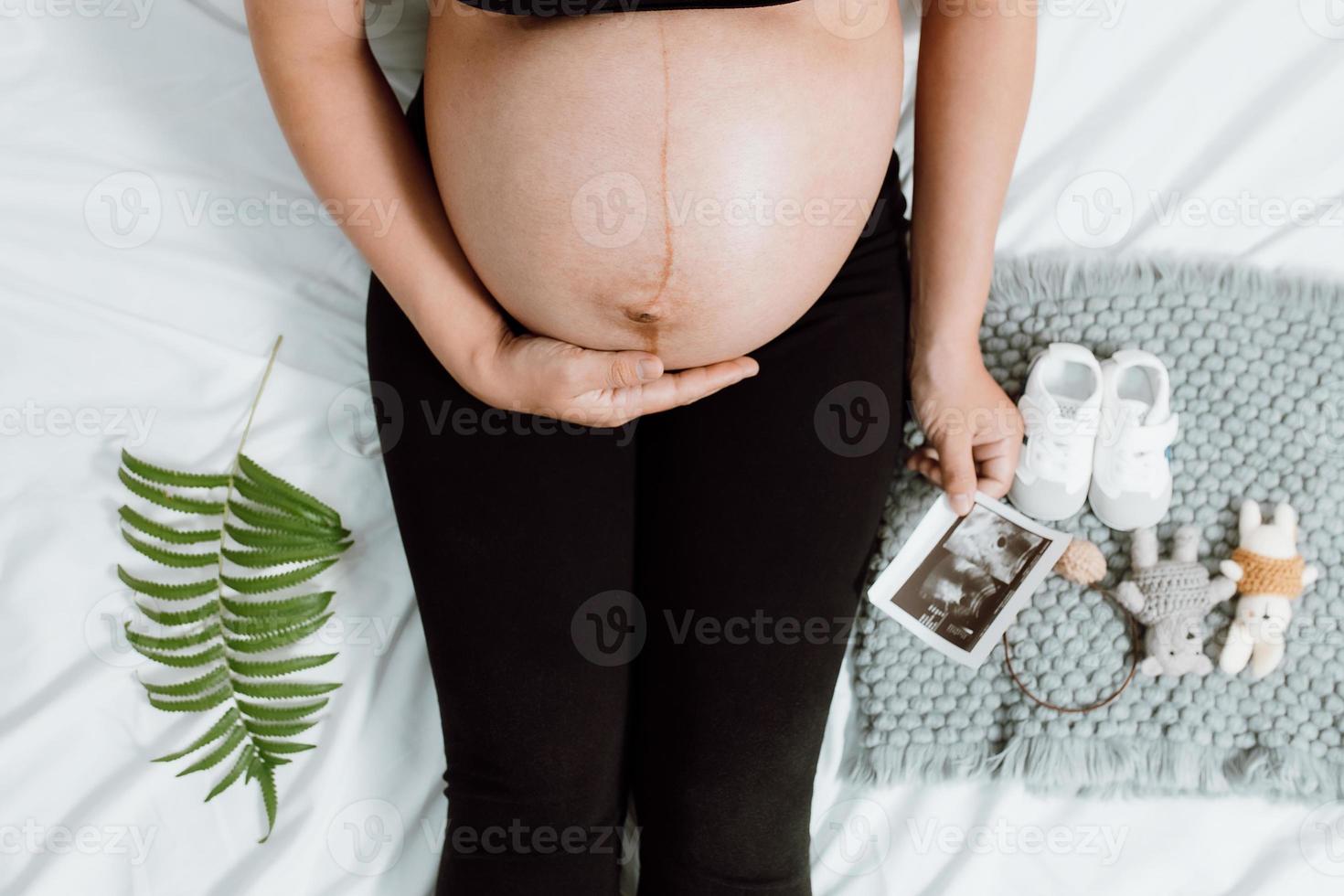 Pregnant woman hands holding on belly making a take care and love symbol on the bedroom with ultrasound image. Expectant mother waiting and preparing for baby birth. Pregnant woman concept. photo