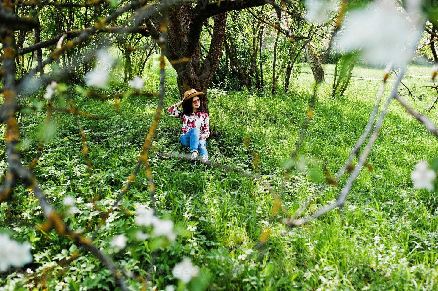 Spring portrait of brunette girl in pink glasses and hat at green blossom garden. photo