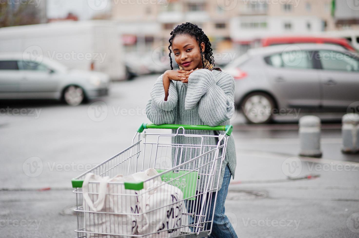 mujer africana con carrito de compras posó en el mercado al aire libre cerca del estacionamiento. foto