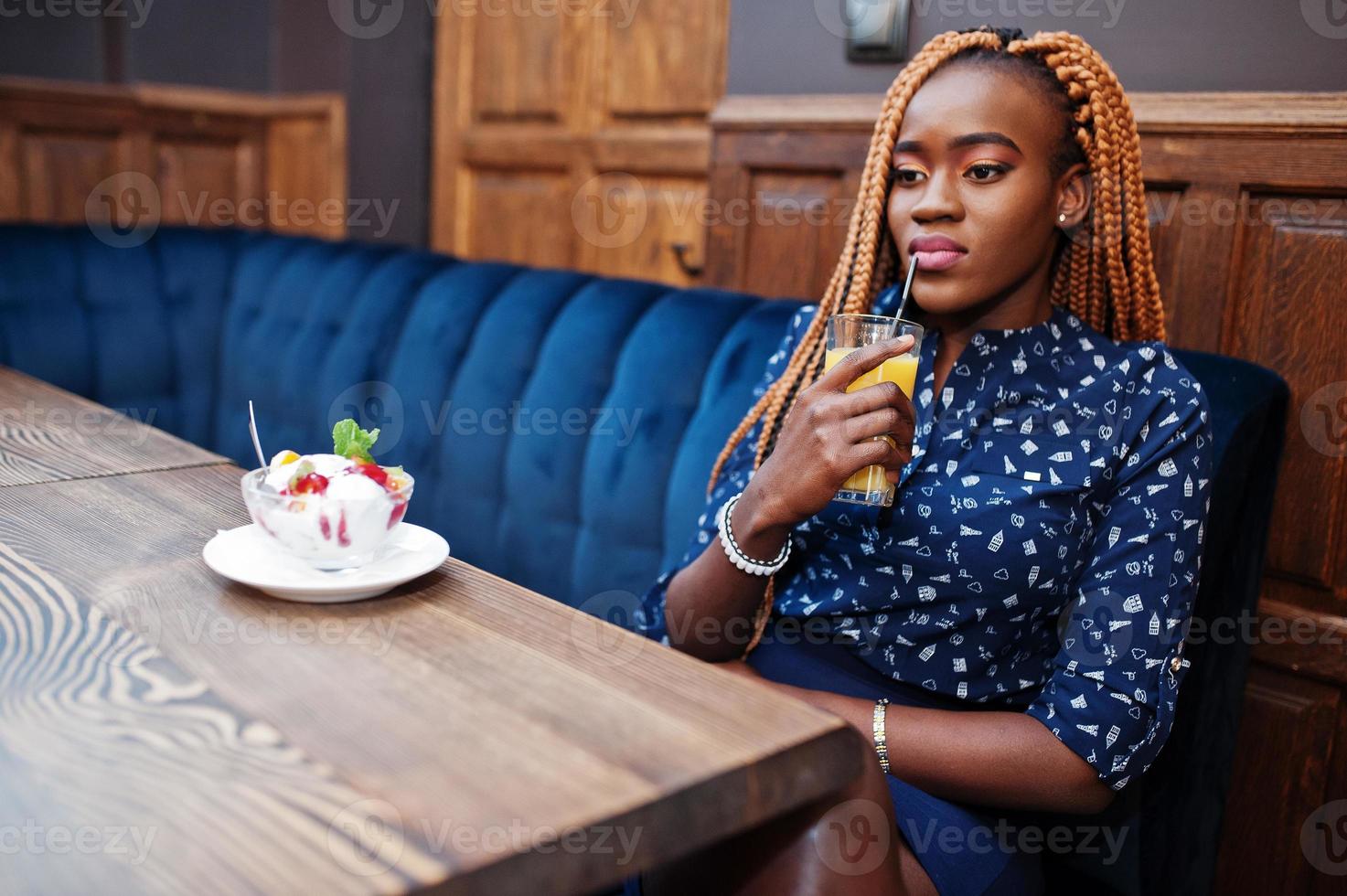 retrato de una hermosa joven mujer de negocios africana con rastas, vestida con blusa y falda azul, sentada en un café con helado y jugo de piña. foto