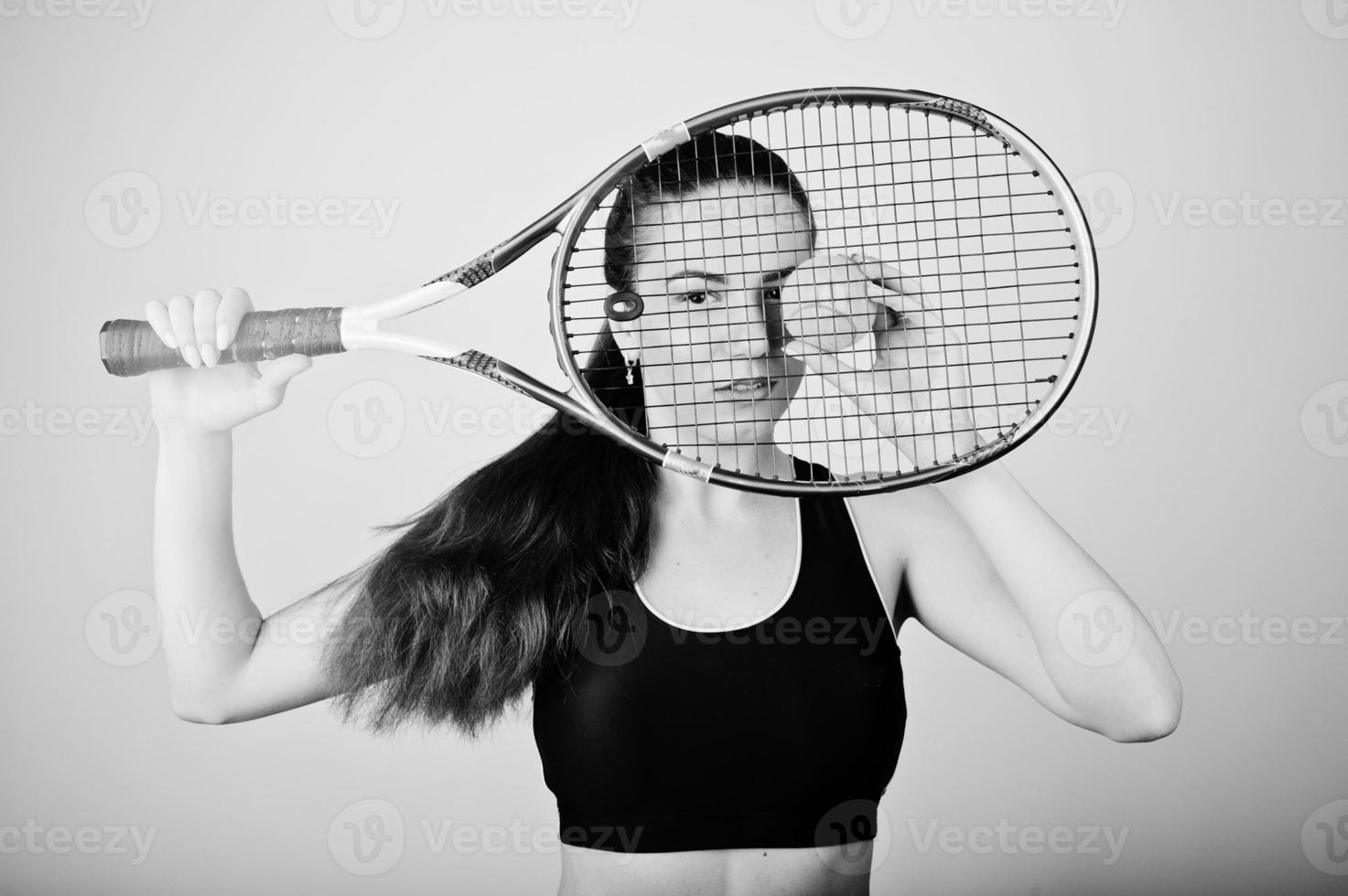 Black and white portrait of beautiful young woman player in sports clothes holding tennis racket while standing against white background. photo