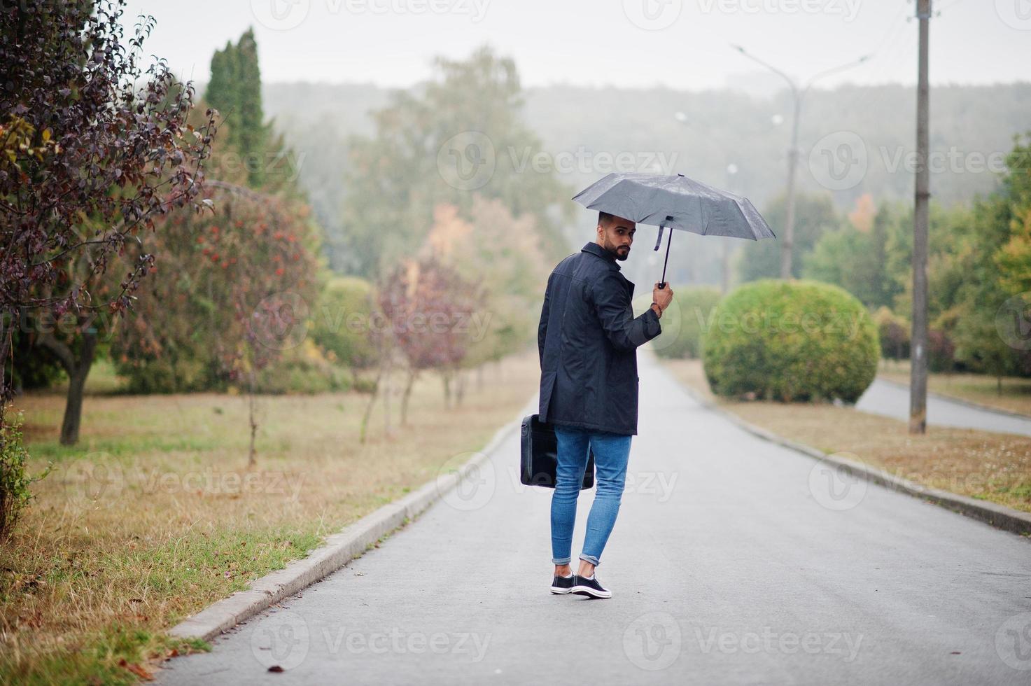 Fashionable tall arab beard man wear on black coat with umbrella and bag case posed at rain weather day. photo