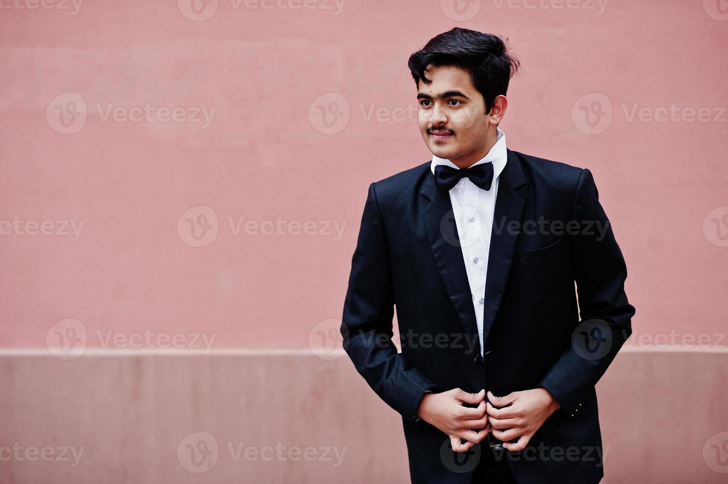 joven indio con traje y corbata de moño posado contra una pared rosa. foto