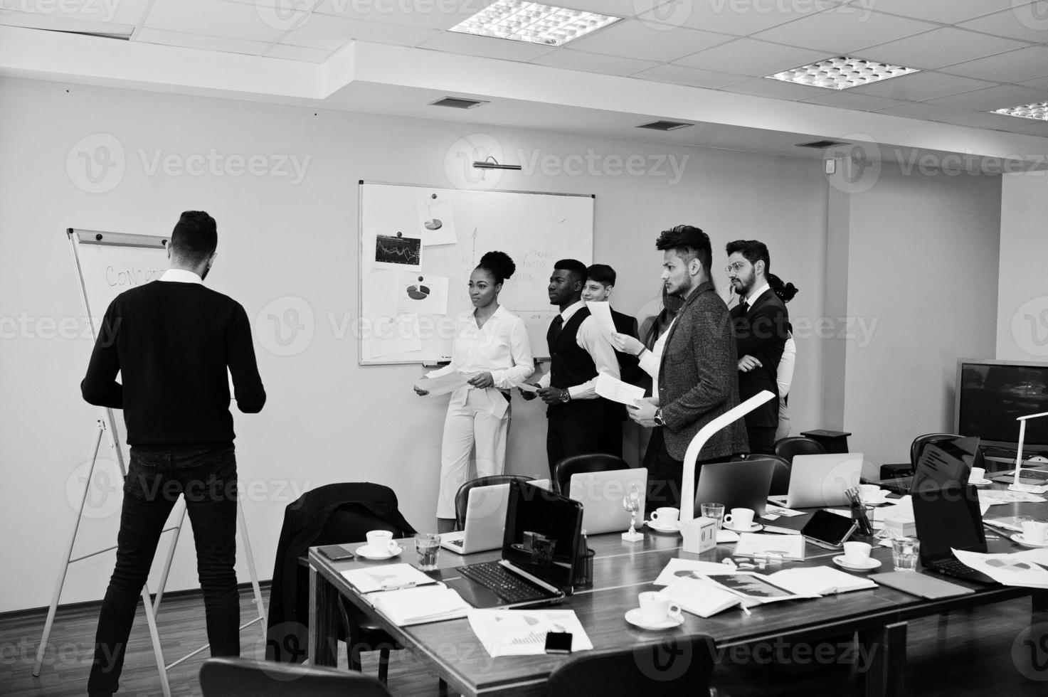 Mixed race business coach presenting report standing near whiteboard pointing on sales statistic shown on diagram and chart teach diverse company members gathered together in conference room. photo