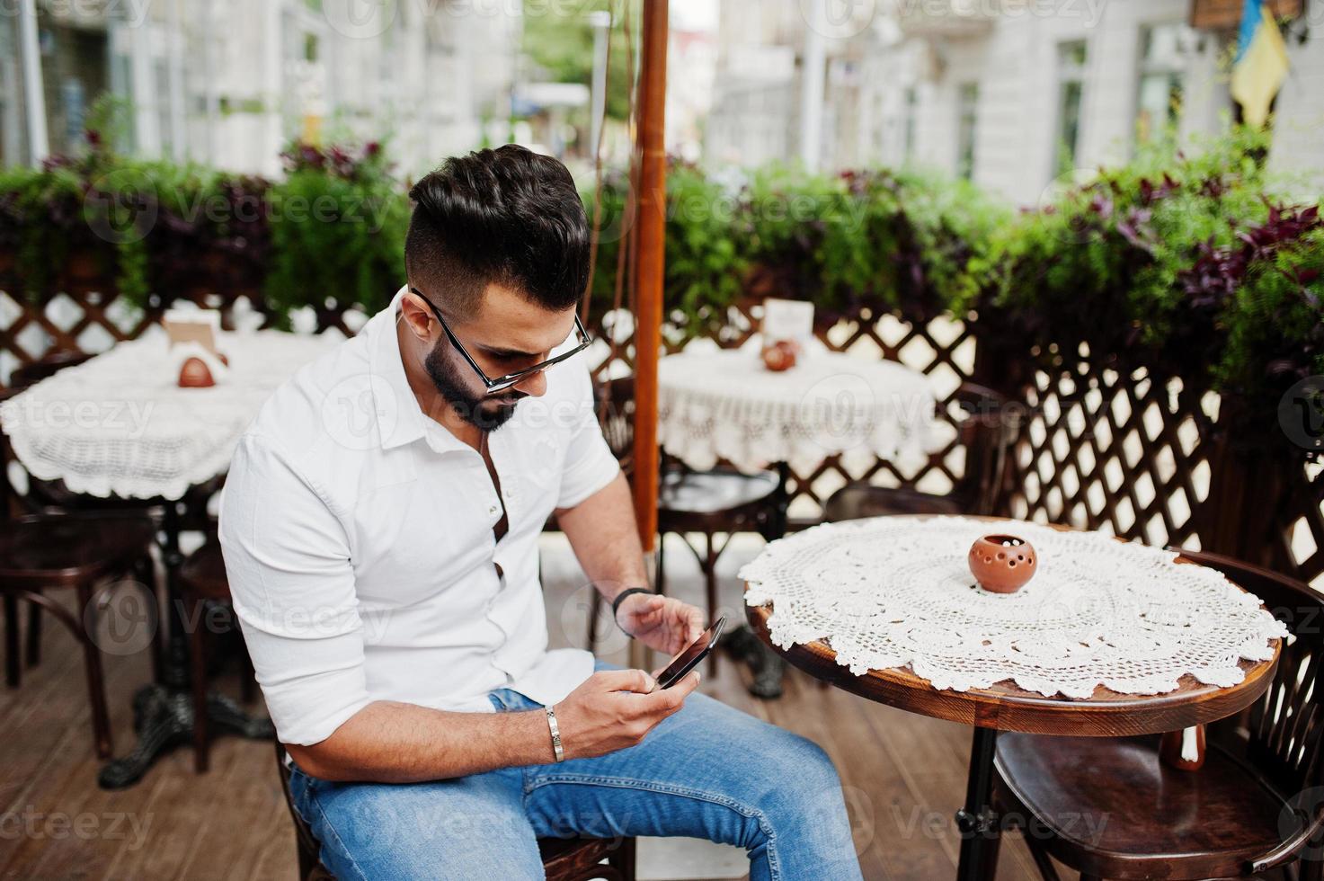 Elegante modelo de hombre árabe alto con camisa blanca, jeans y gafas de sol en la calle de la ciudad. Barba atractivo chico árabe sentado en un café al aire libre y mirando el teléfono móvil. foto