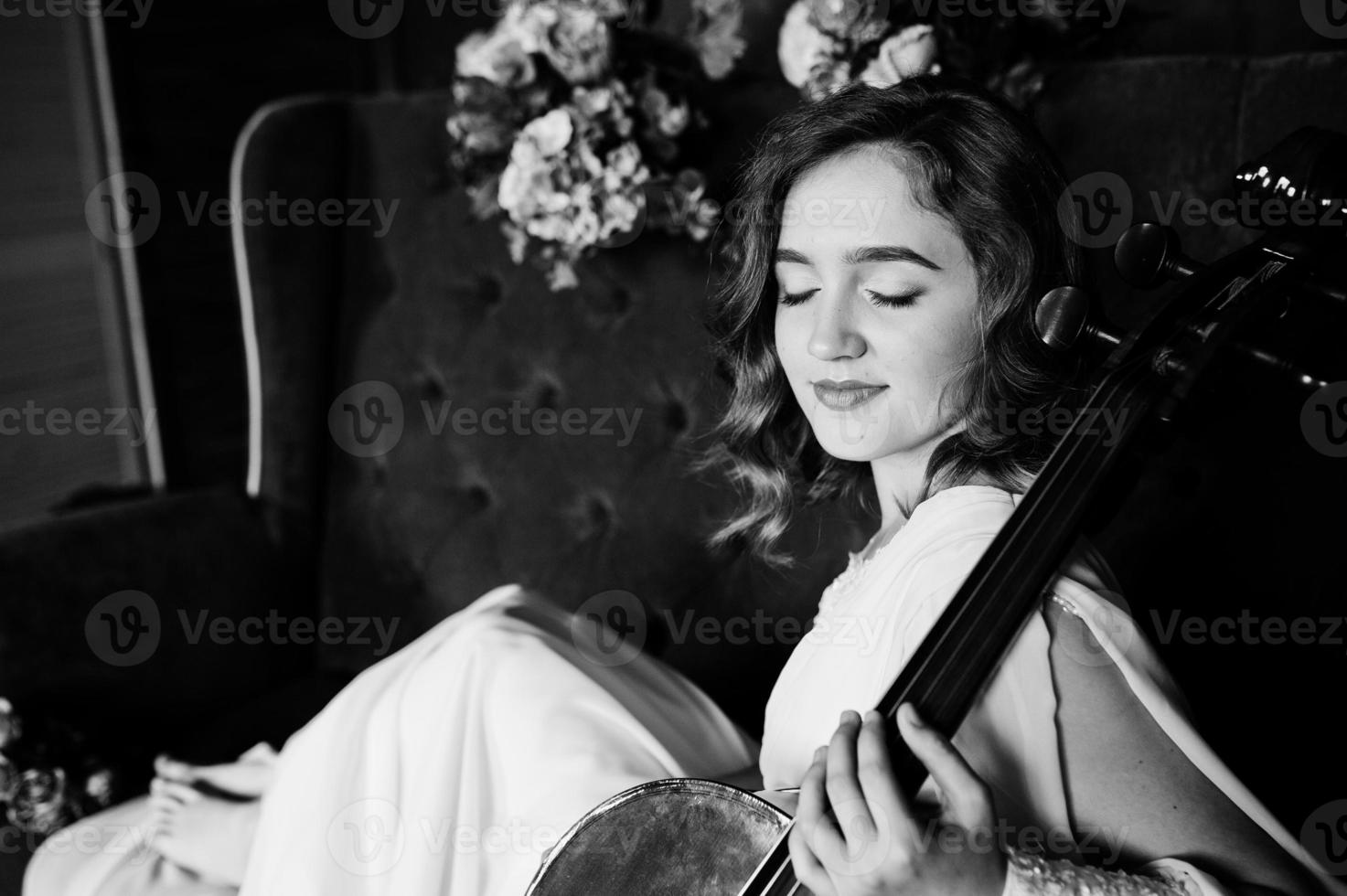 bastante joven músico gilrl en vestido blanco con contrabajo sentado en un sofá vintage marrón. foto