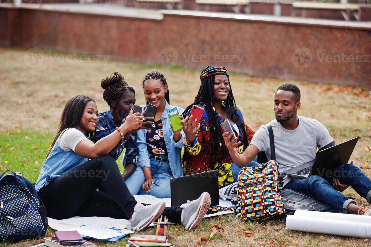 Group of five african college students spending time together on campus at university yard. Black afro friends making with mobile phones. Education theme. photo