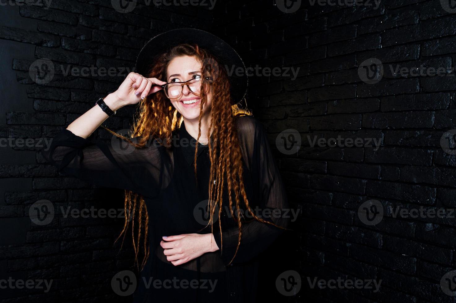 sesión de estudio de una chica de negro con rastas, anteojos y sombrero sobre fondo de ladrillo. foto