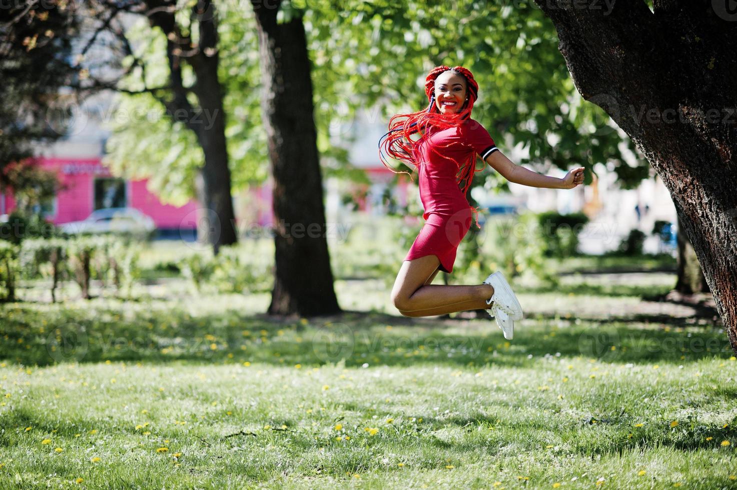 Cute and slim african american girl in red dress with dreadlocks jump outdoor in spring park. Stylish black model. photo