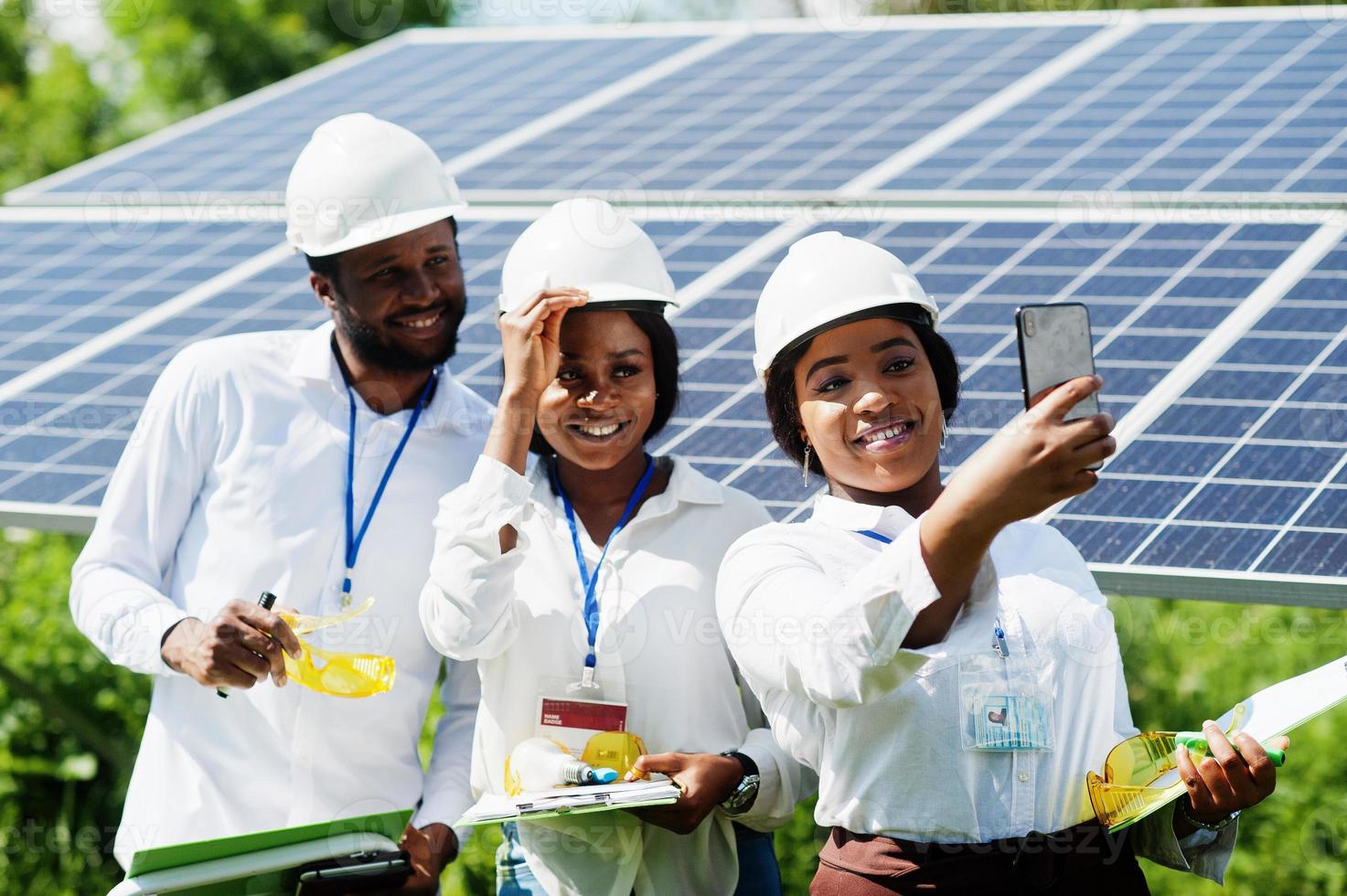 técnico afroamericano comprueba el mantenimiento de los paneles solares. grupo de tres ingenieros negros reunidos en la estación solar. hacer selfie por teléfono. foto