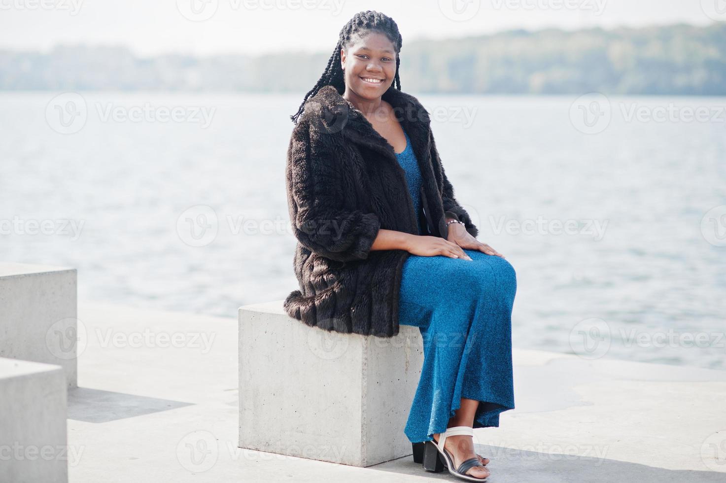 modelo afroamericano de piel oscura de talla grande posado con un vestido azul brillante y un abrigo de piel negro sentado en un cubo de piedra frente al mar. foto