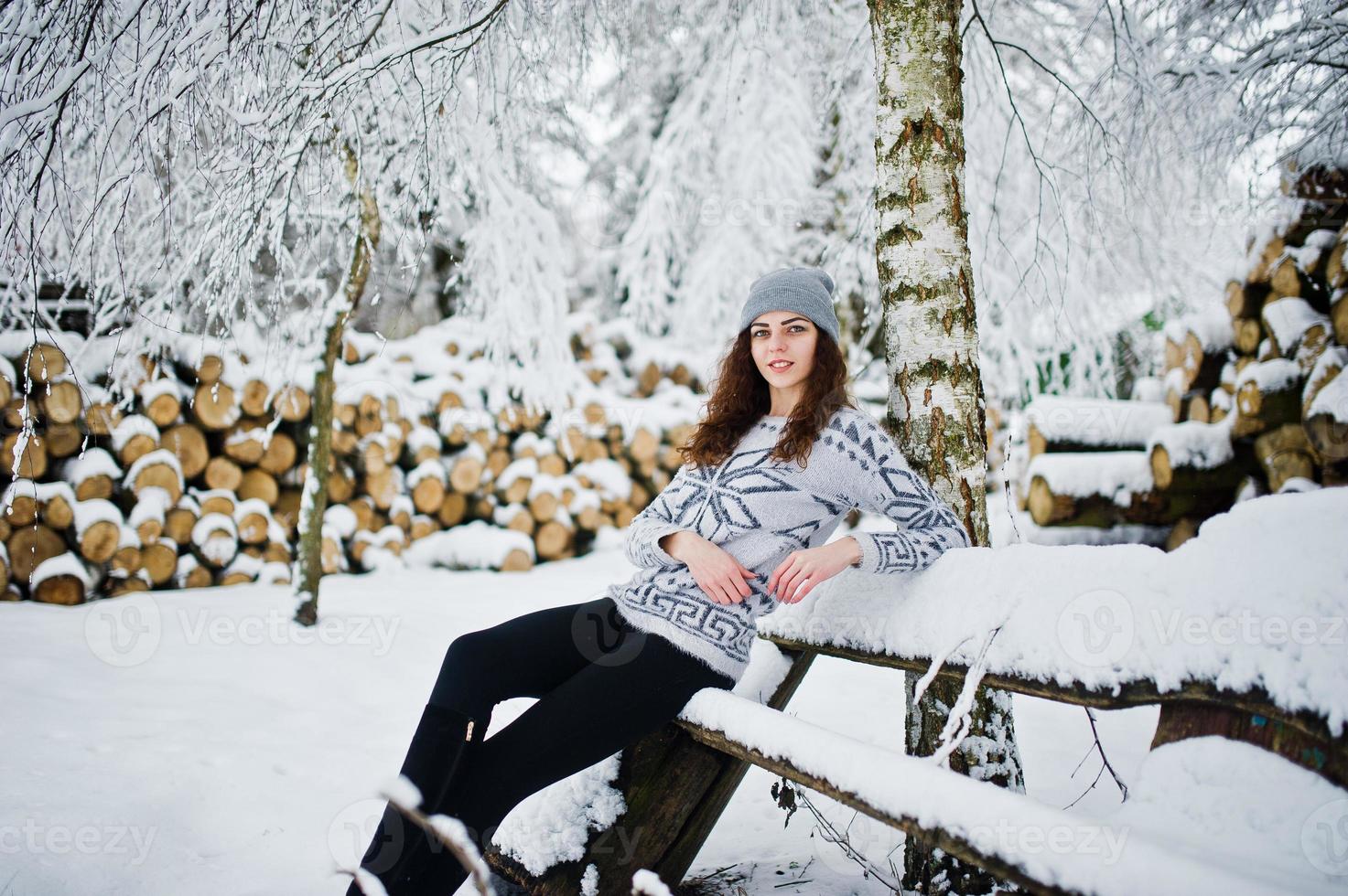 linda chica rizada con suéter y sombreros sentada en un banco en el parque forestal nevado en invierno. foto