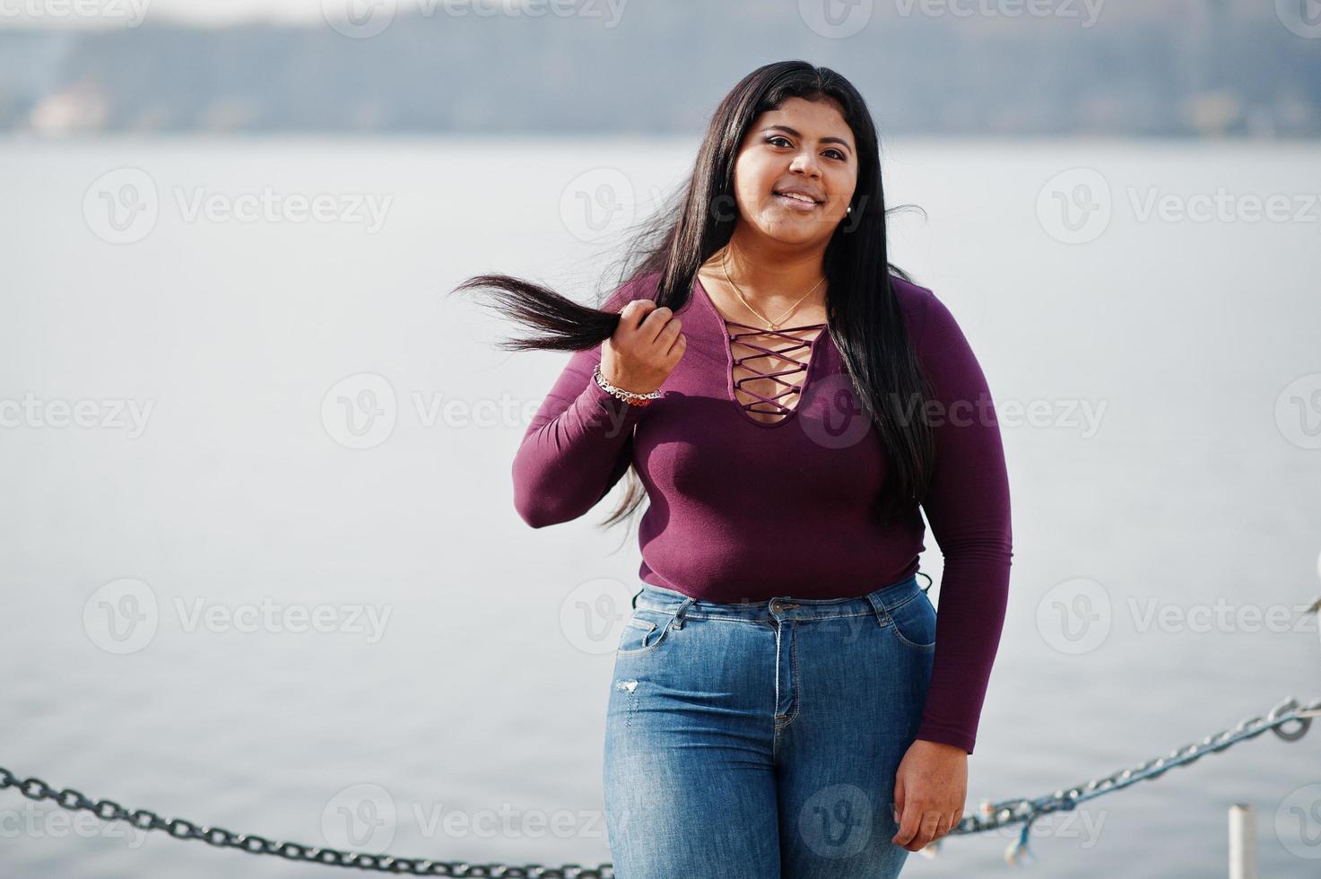 Pretty latino xxl model girl from Ecuador wear on violet blouse posed against lake. photo