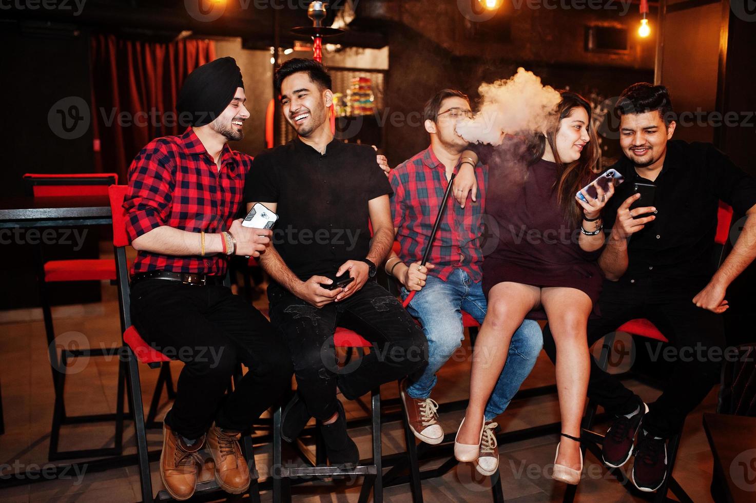 Group of indian friends sitting at lounge bar, having fun and rest, smoking hookah and looking on mobile phones. photo