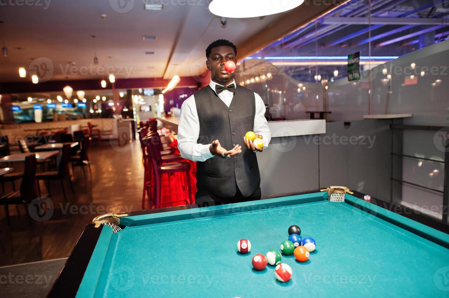 joven apuesto hombre africano vestido con camisa blanca, chaleco negro y corbata de moño jugar al billar. foto