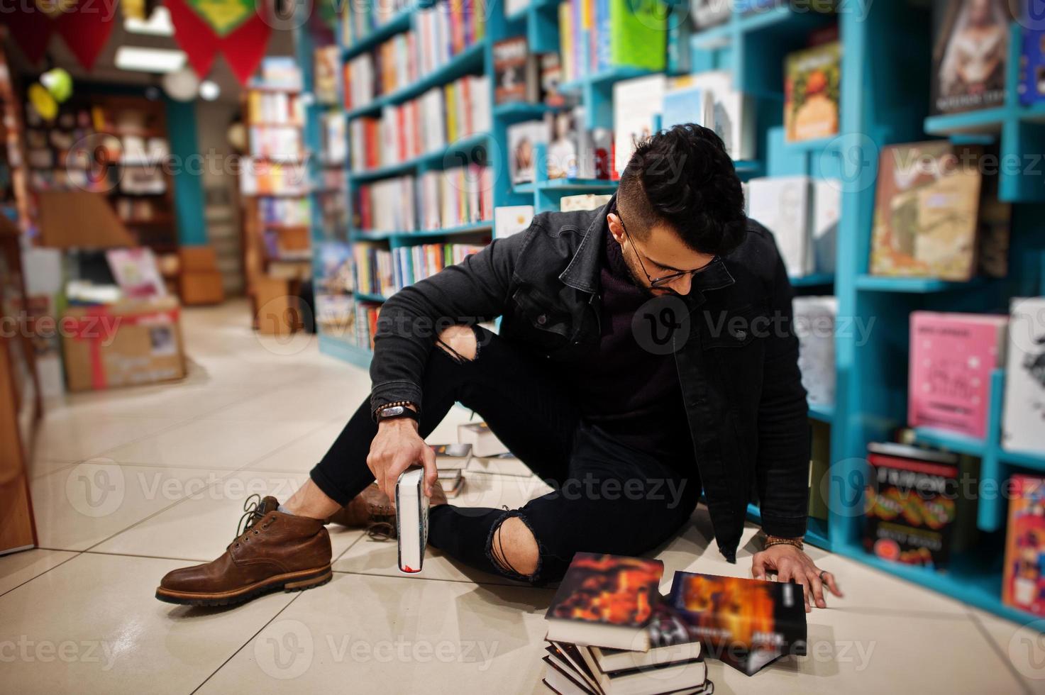 estudiante árabe alto e inteligente, vestido con chaqueta negra de jeans y anteojos, en la biblioteca sentado contra estantes de libros. foto