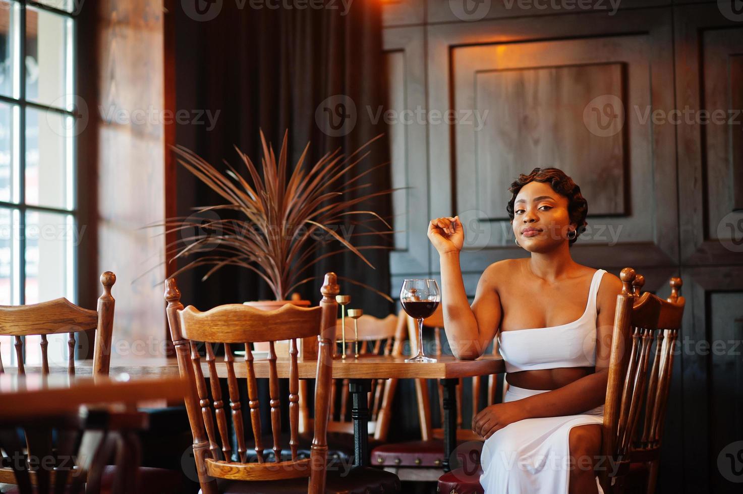 mujer afroamericana, peinado retro con vestido blanco en el restaurante con una copa de vino. foto