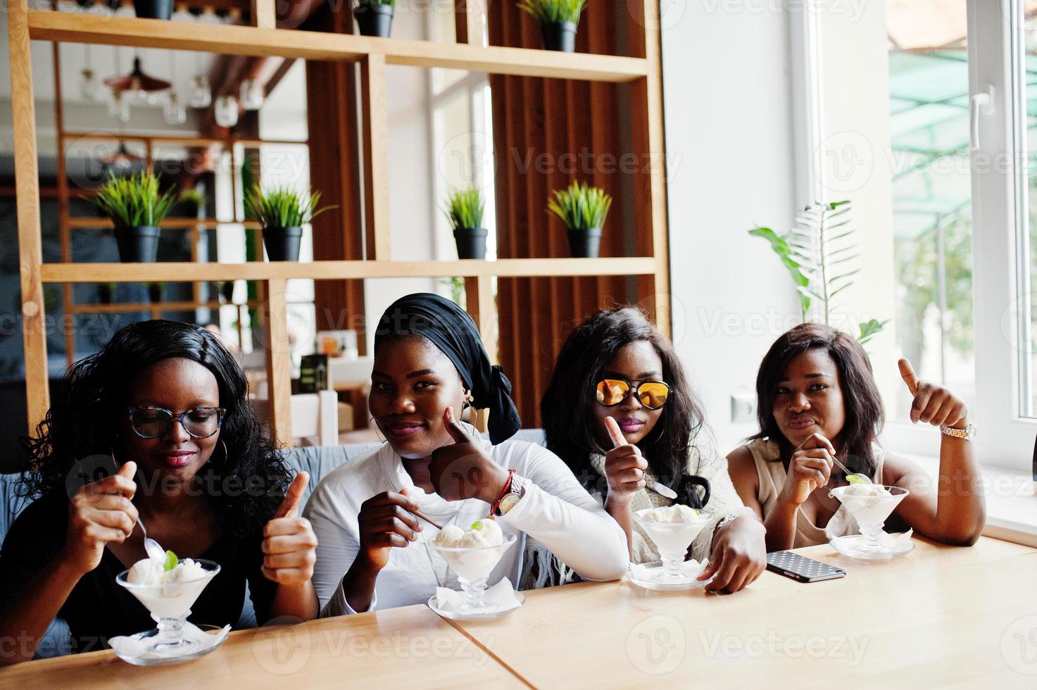 Four african american girls sitting on table at cafe and eating ice cream dessert and showing thumb up. photo