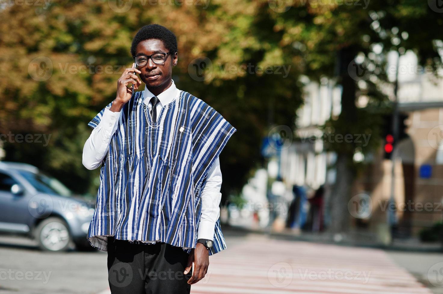 African man in traditional clothes and glasses walking at crosswalk and speaking on mobile phone. photo