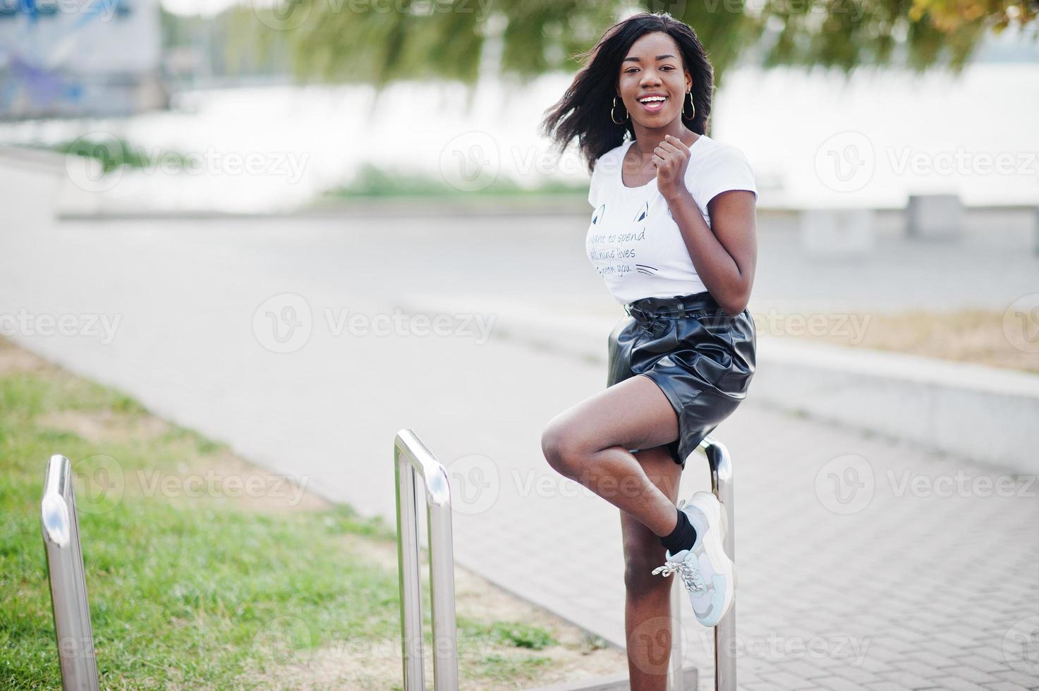 Modelo delgado afroamericano de piel oscura posado en pantalones cortos de cuero negro y camiseta blanca. foto