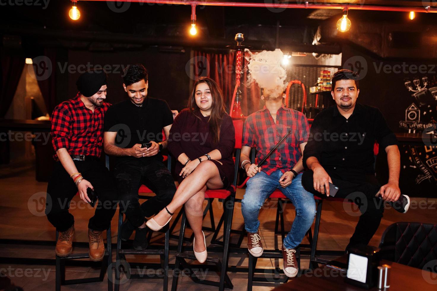 Group of indian friends sitting at lounge bar, having fun and rest, smoking hookah and looking on mobile phones. photo