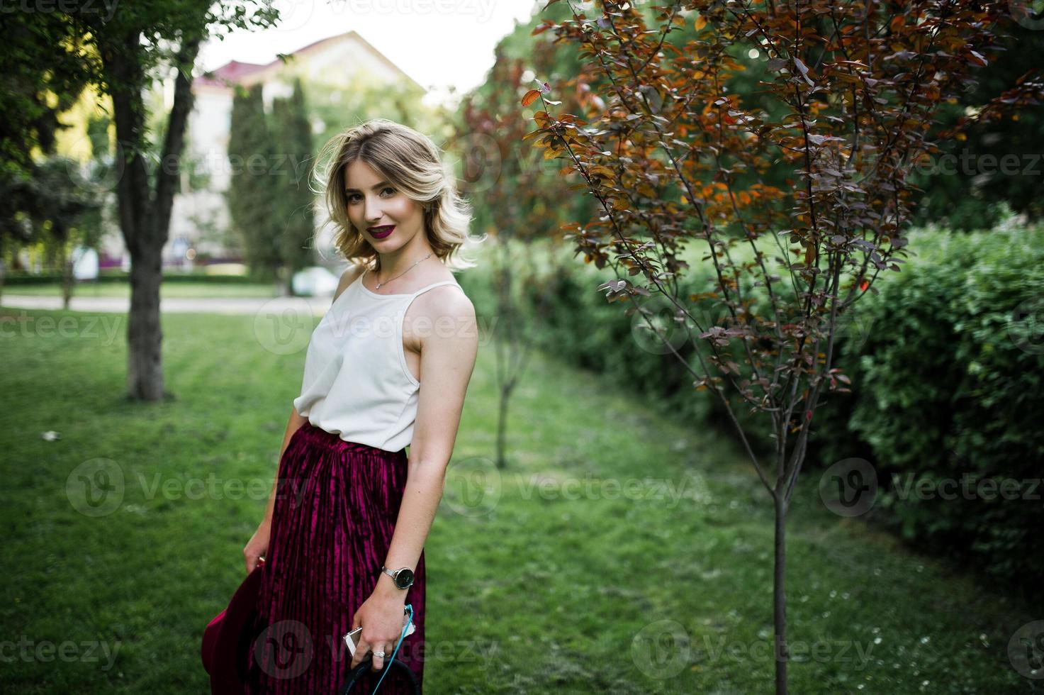 Fashionable and beautiful blonde model girl in stylish red velvet velour skirt, white blouse and hat, posed at park with phone and earphones. photo