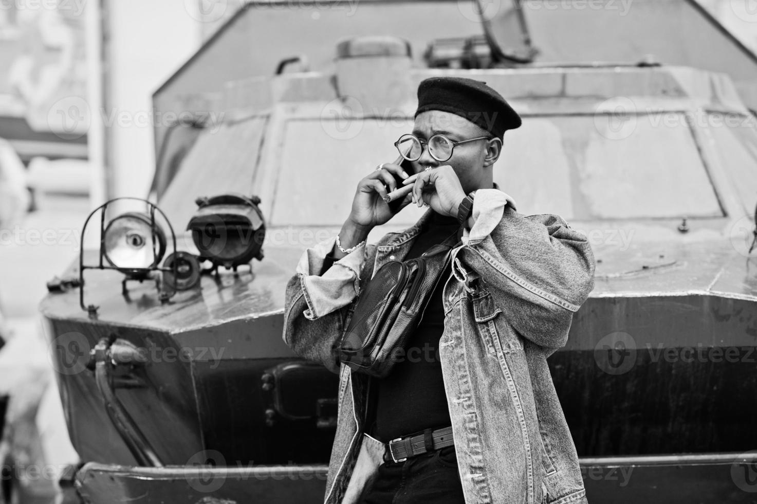 African american man in jeans jacket, beret and eyeglasses, with cigar posed against btr military armored vehicle, and speaking on mobile phone. photo