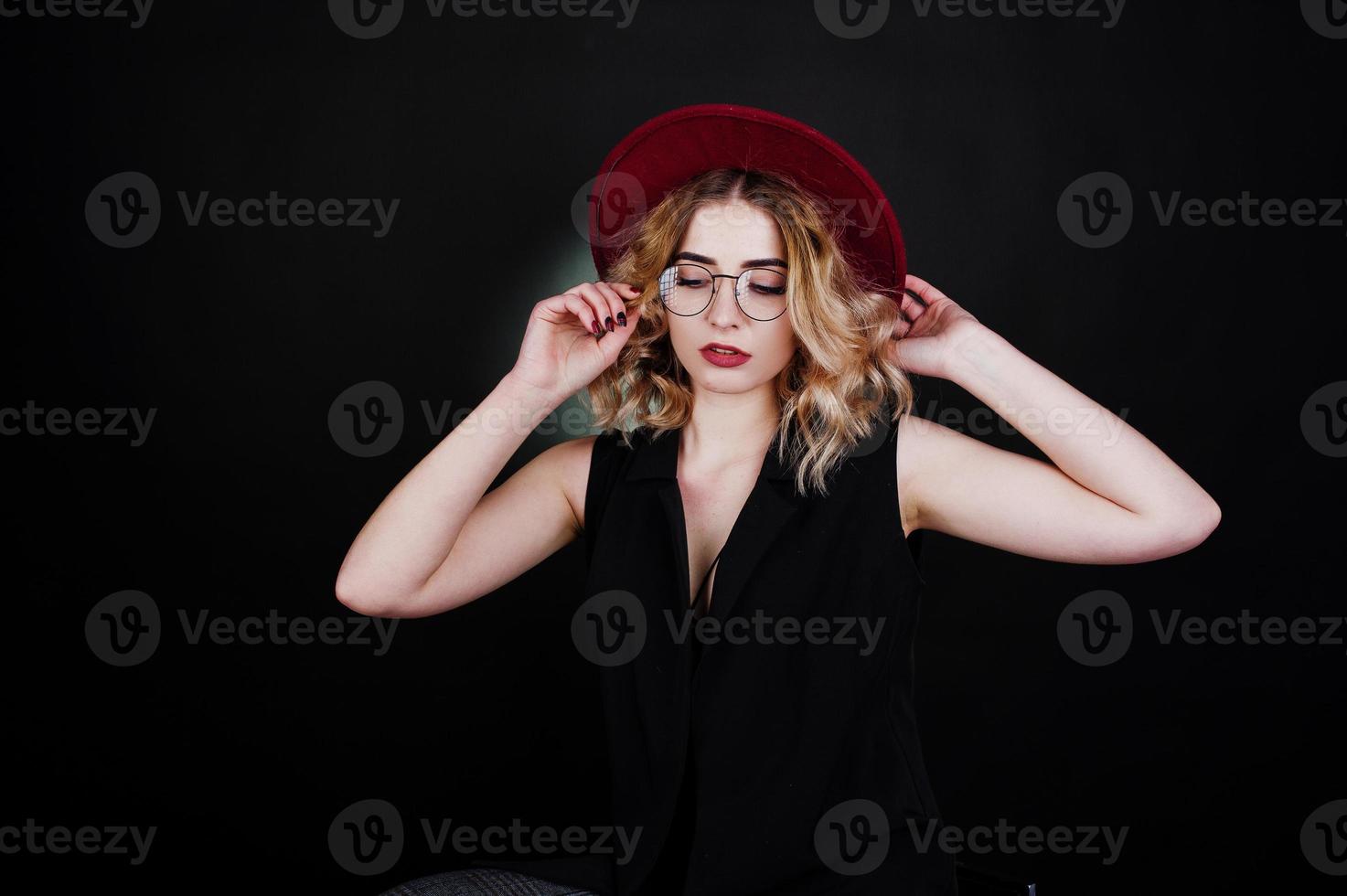 retrato de estudio de una chica rubia con ropa negra, sombrero rojo y anteojos contra un fondo oscuro. foto