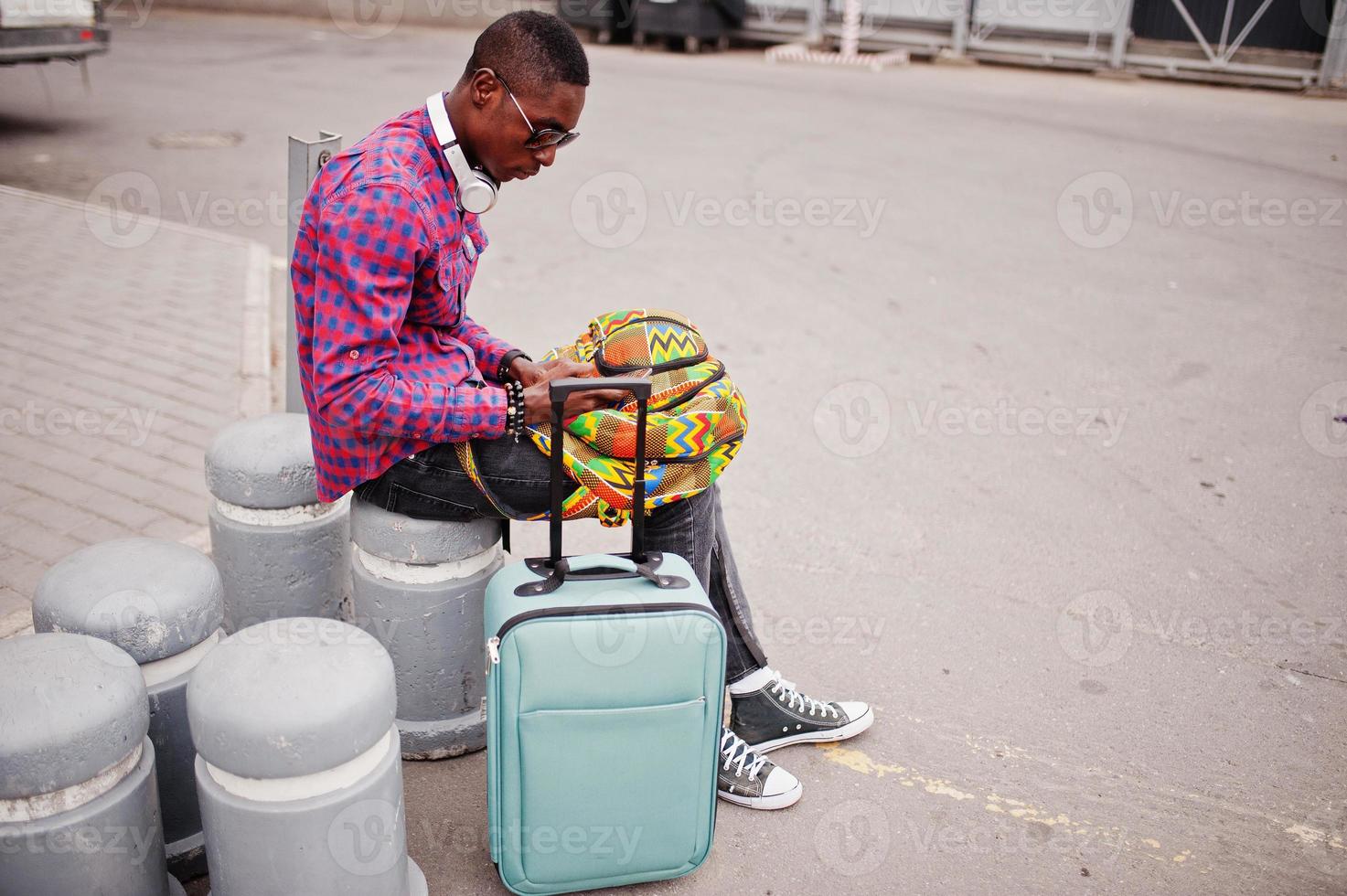 African american man in checkered shirt, sunglasses and earphones with suitcase and backpack. Black man traveler holding mobile phone at hand. photo