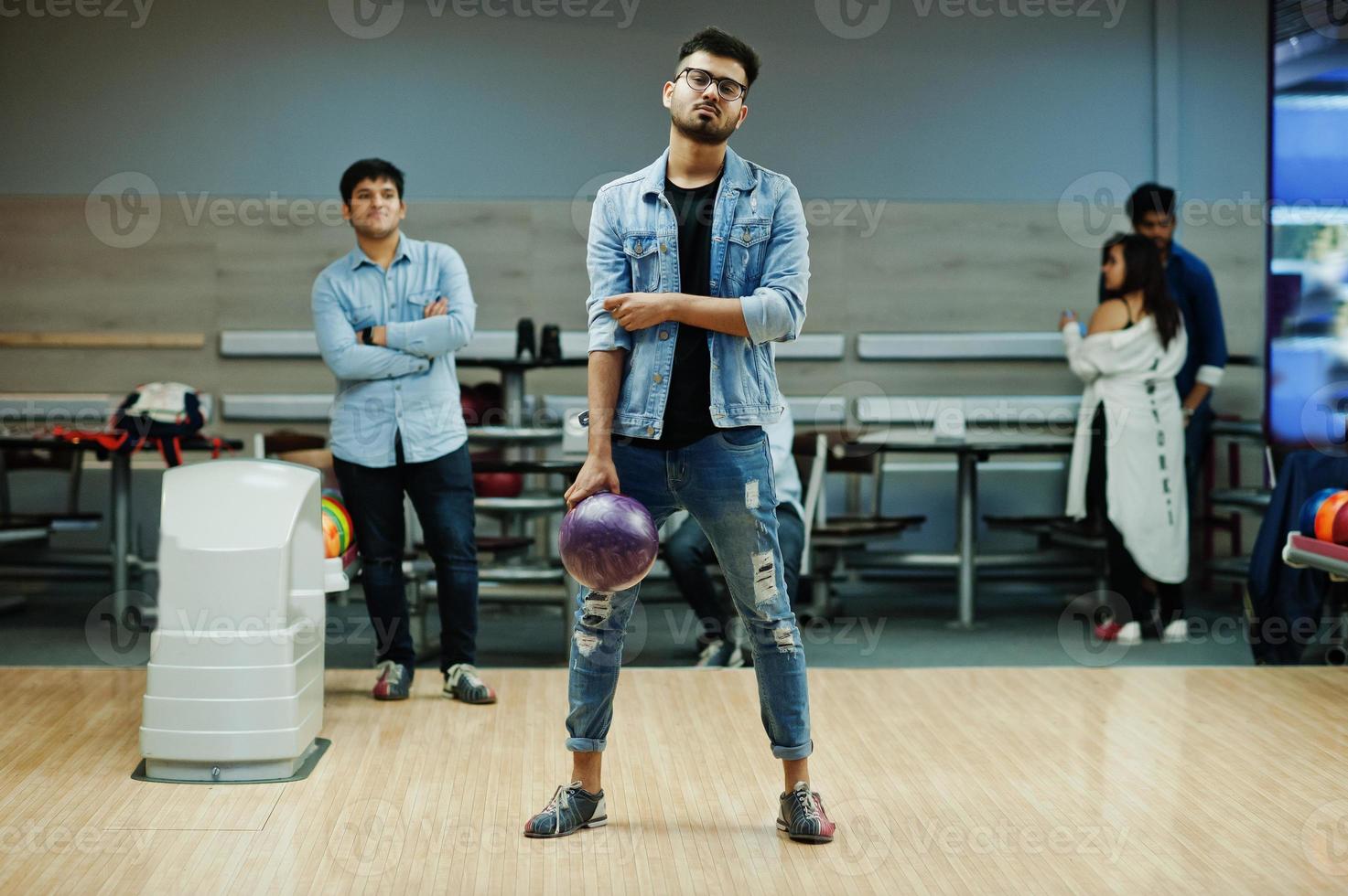 Stylish asian man in jeans jacket and glasses standing at bowling alley with ball at hand. photo