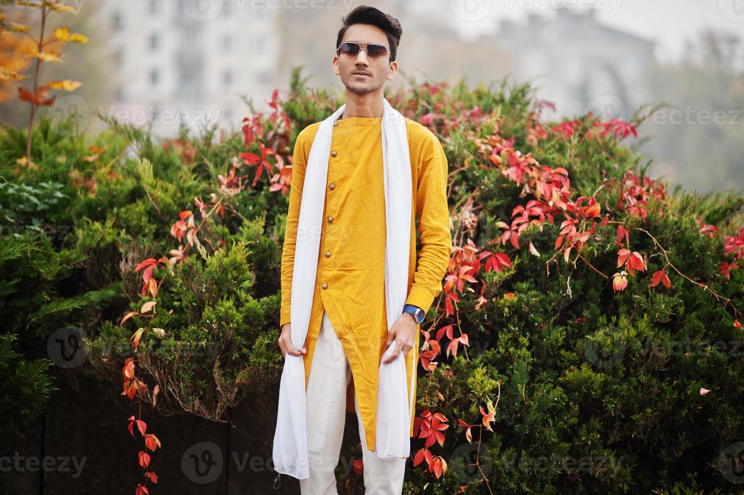 Indian stylish man in yellow traditional clothes with white scarf, sunglasses posed outdoor against autumn leaves tree. photo