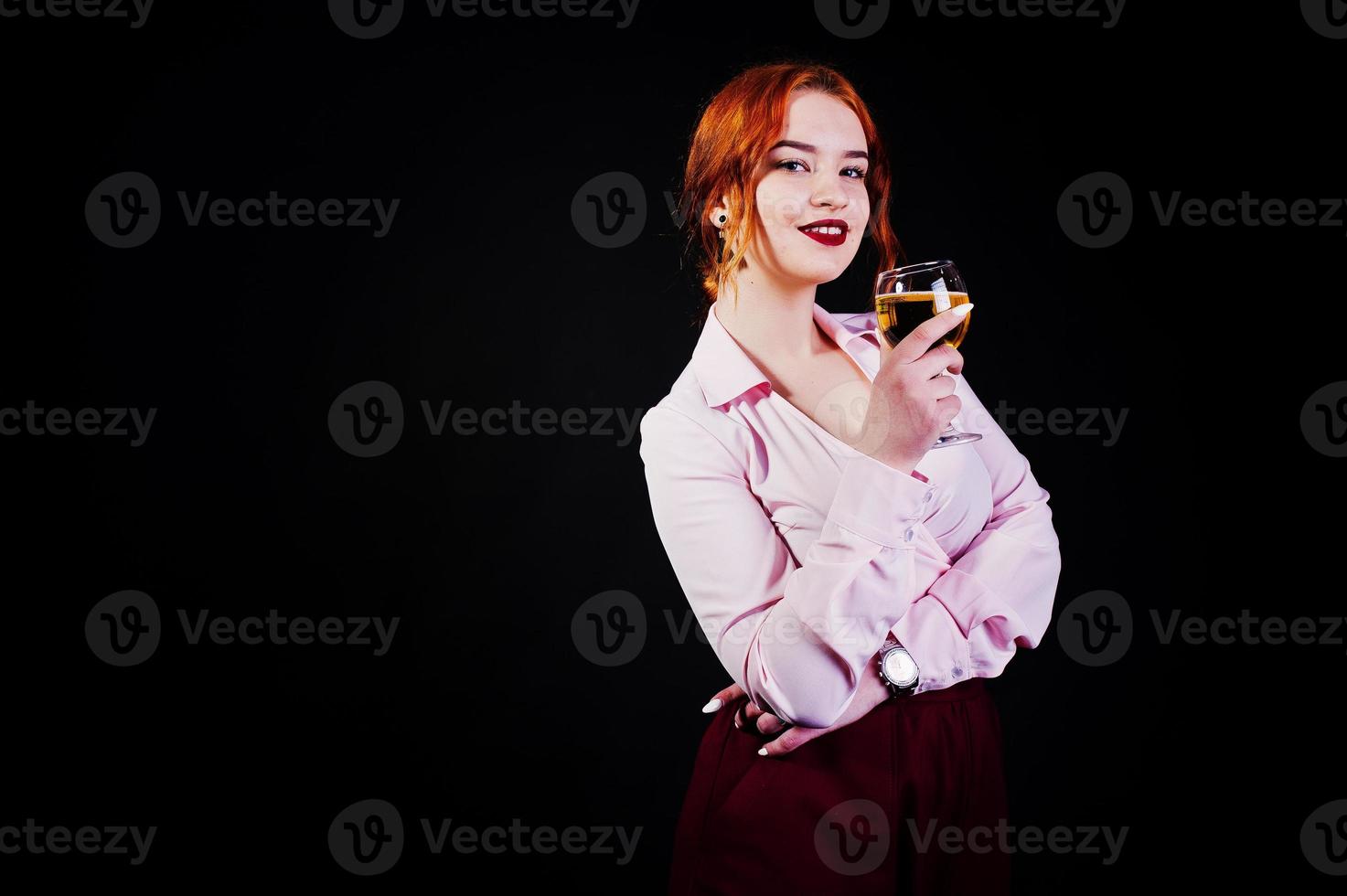 Gorgeous red haired girl in pink blouse and red skirt with glass of wine at hand isolated on black. photo
