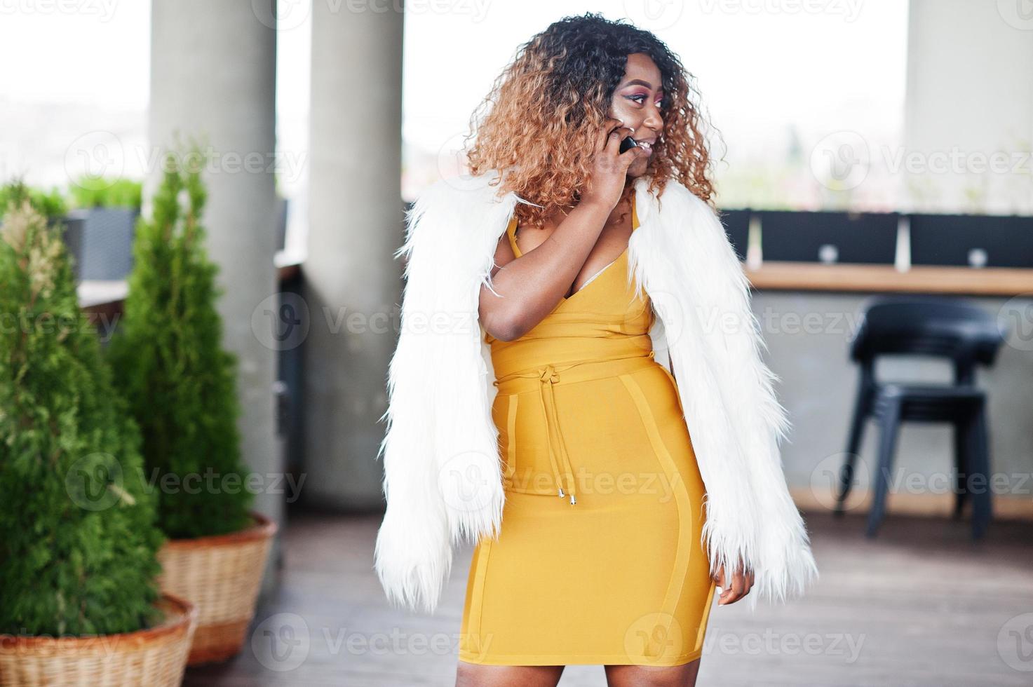 Glamour african american woman in yellow dress and white woolen cape posed at balcony and speak on mobile phone. photo