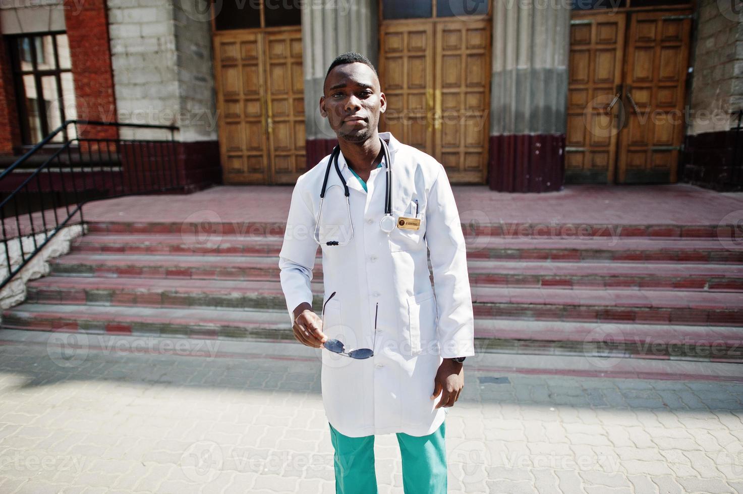 African american doctor male at lab coat with stethoscope outdoor against clinic door. photo