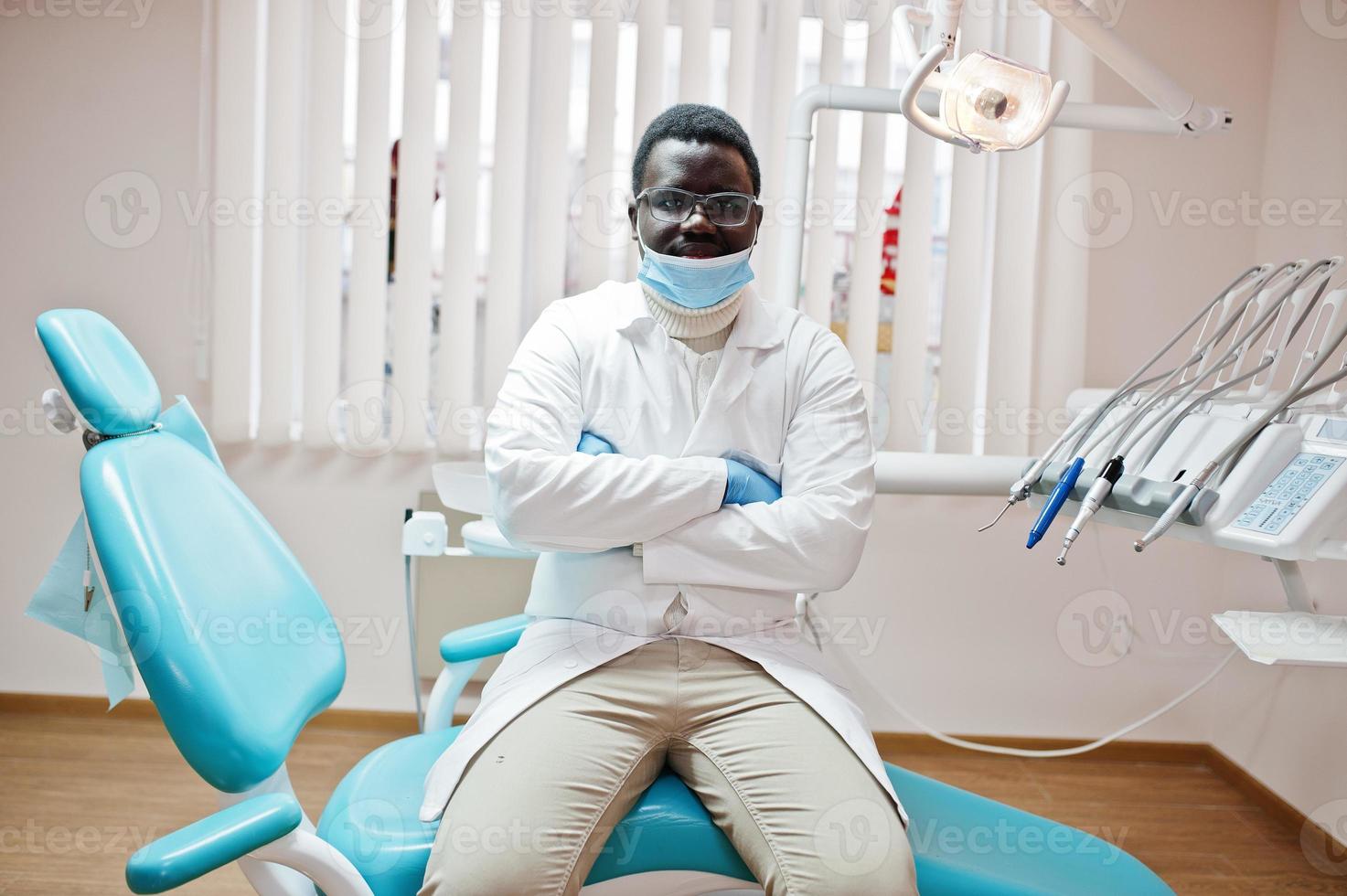 médico afroamericano con mascarilla y anteojos con los brazos cruzados sentado en la silla del dentista en la clínica dental. foto